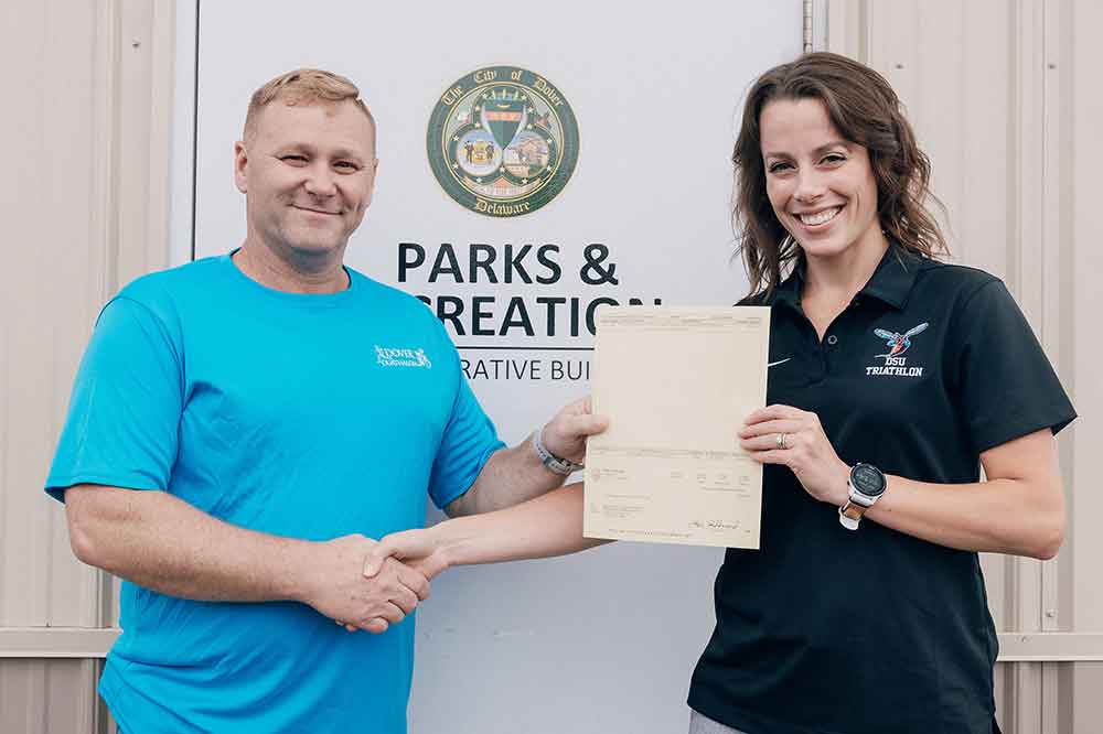 Robin Eaton (l), director of the Dover Department of Parks and Recreation, presents a $2,000 check to University Triathlon Head Coach Hannah P. Loftus for her team's work in assisting in the running of the city's June 22 Duathlon event.