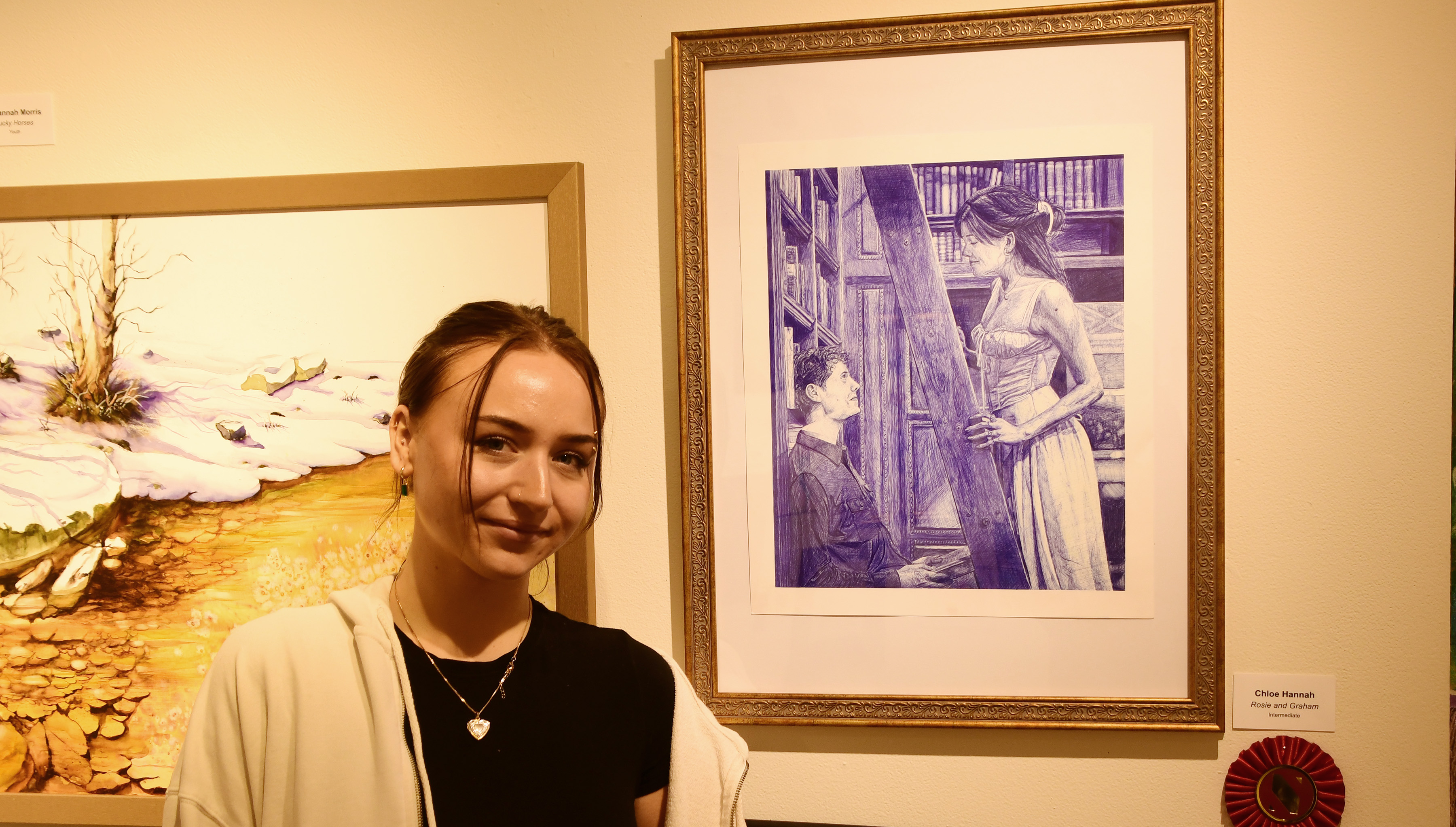 Chloe Hannah, a junior Student Art major, stands next to her ball point pen work "Rosie and Graham" which won 2nd Place in the Intermediate Category of the State Employees Art Exhibition. Ms. Hannah is the daughter of a state employee, immediate family members of which were permitted to submit works.